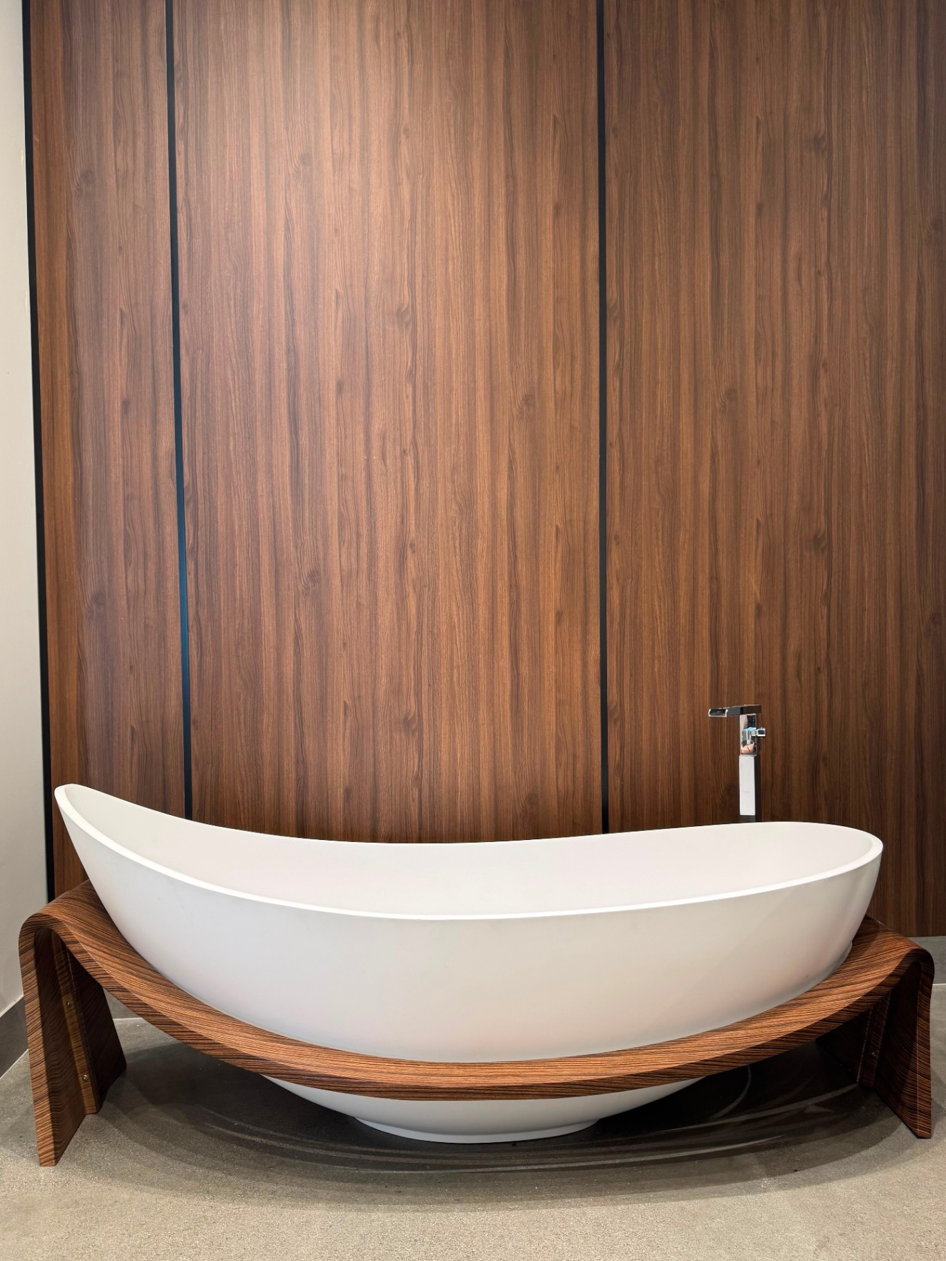 Modern white freestanding slipper bathtub on curved walnut wood cradle with floor-mounted chrome faucet at Ward Design Center bathroom display