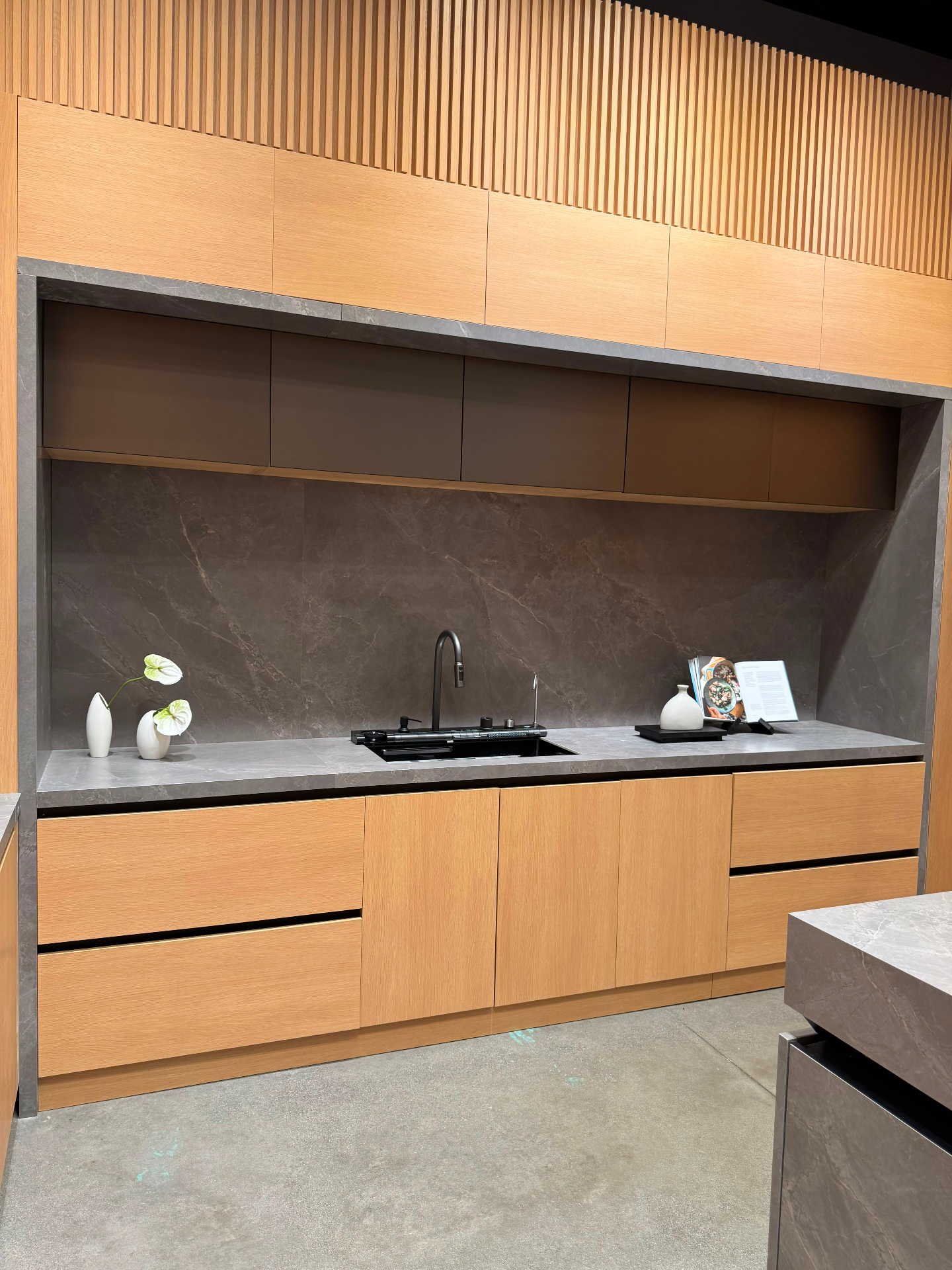 Light oak handleless kitchen cabinetry with grey porcelain stone backsplash, integrated smart sink and matte upper cabinets at Ward Design Center Kapolei