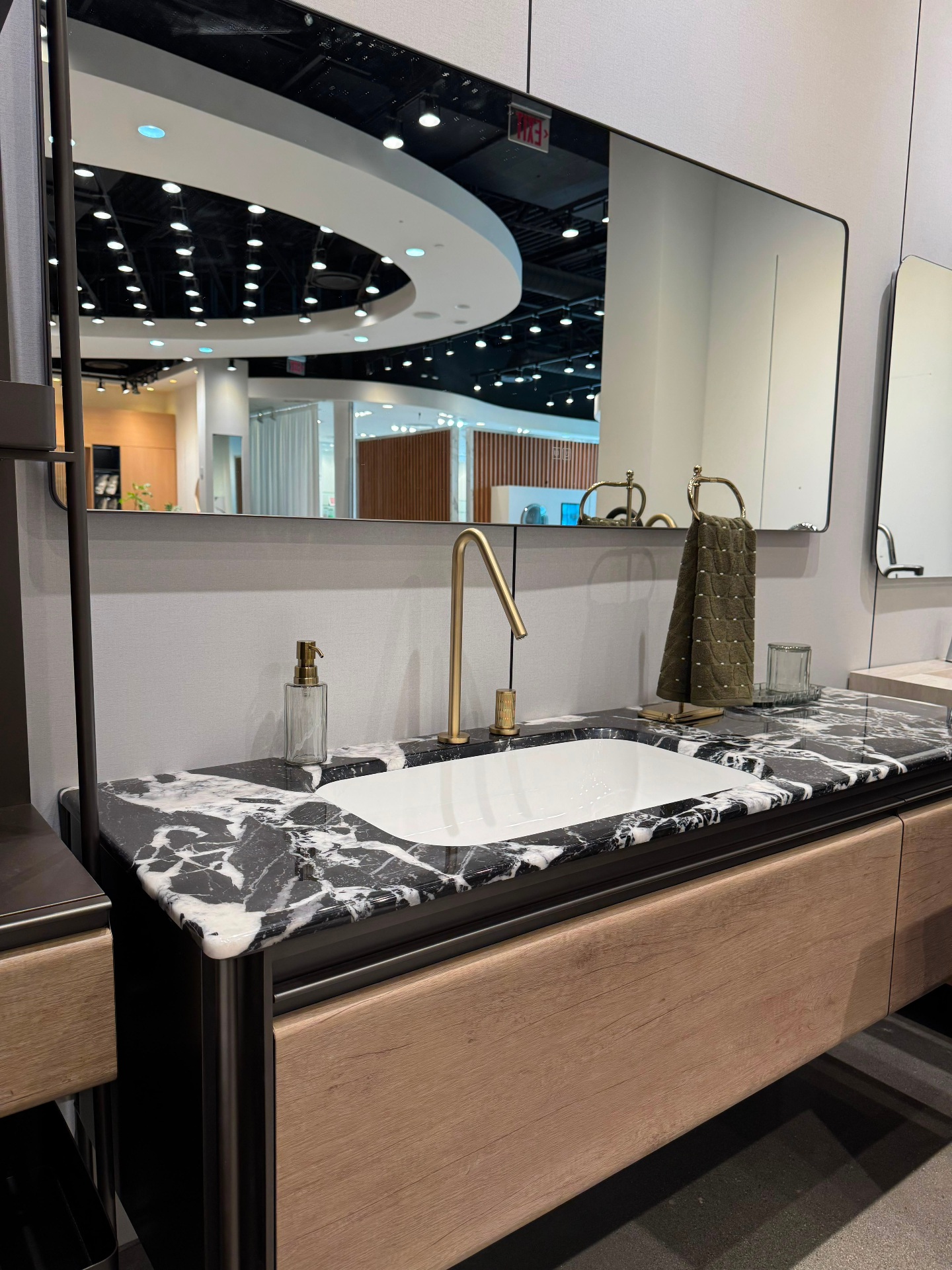 Wall-mounted oak vanity with dramatic black marble countertop, undermount sink and brushed brass tall basin faucet — Ward Design Center Kapolei bathroom display