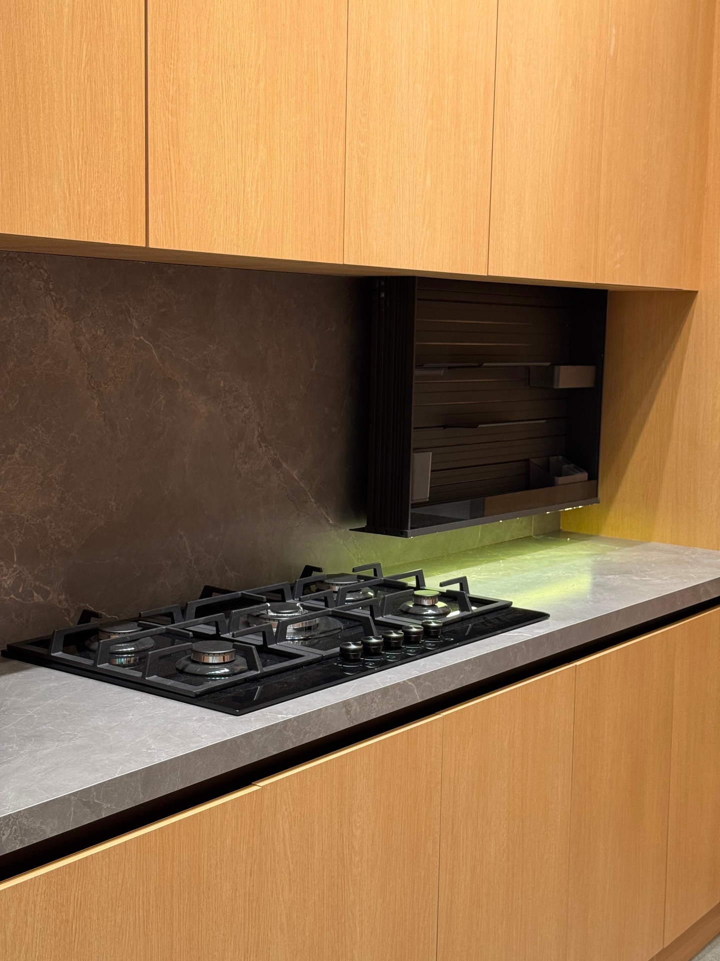 Black 5-burner gas cooktop with porcelain stone backsplash and motorized pull-down upper cabinet — Ward Design Center kitchen display