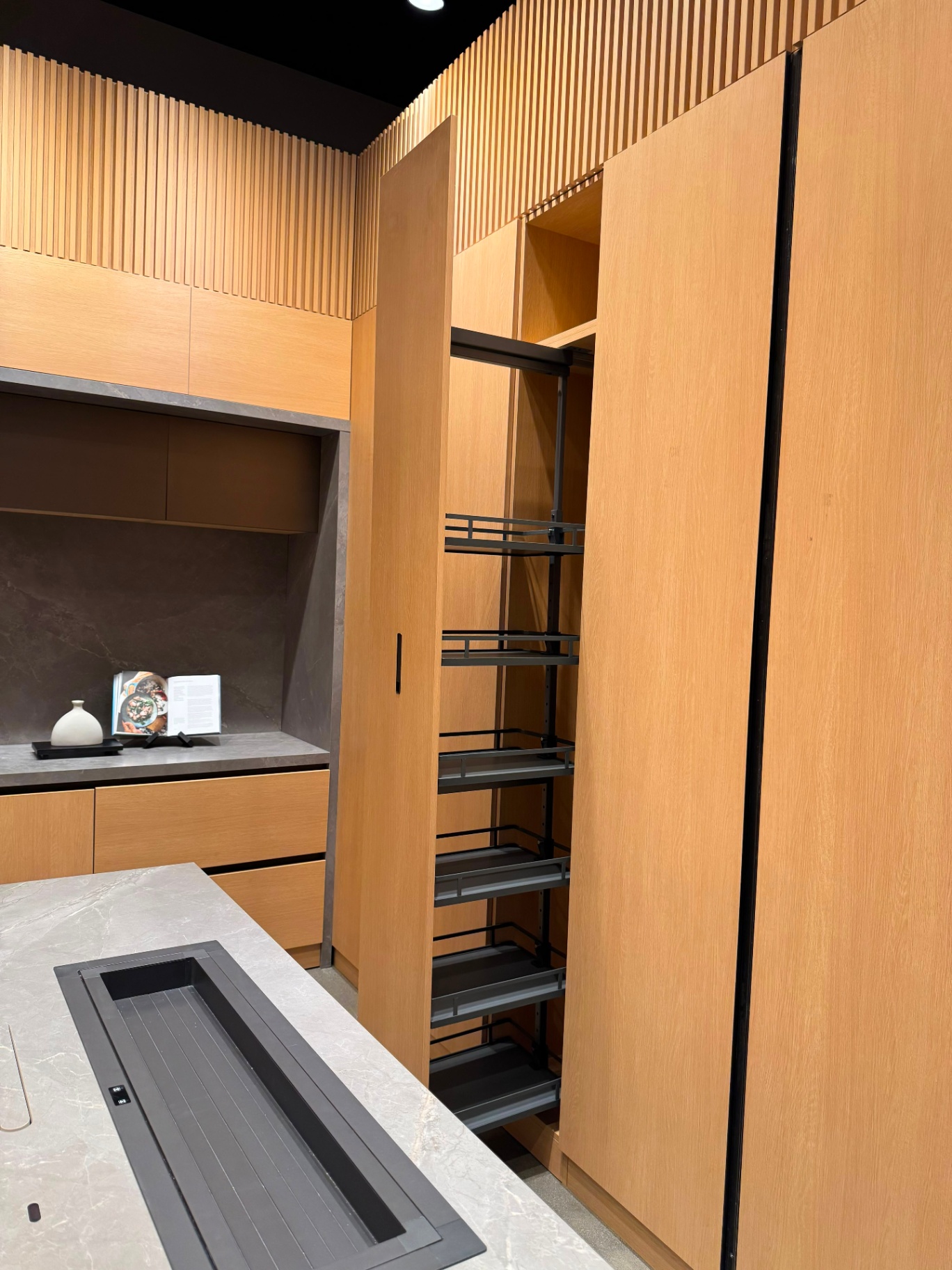 Modern oak kitchen display with porcelain stone countertop, downdraft cooktop, and full-height pantry pull-out at Ward Design Center Kapolei showroom