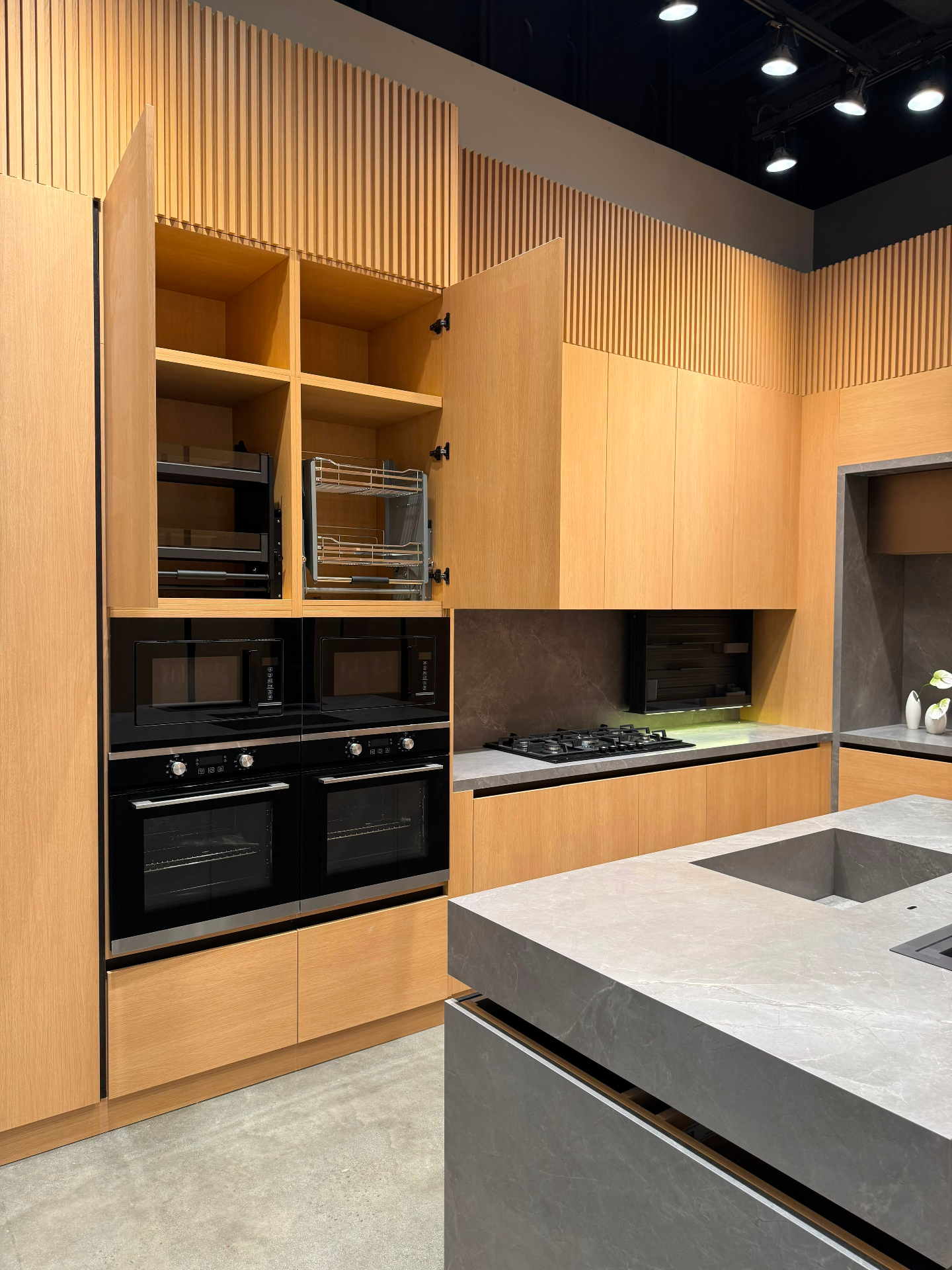 Oak kitchen display with built-in double wall ovens, 4-burner gas cooktop, vertical slat ceiling detail and porcelain stone island at Ward Design Center showroom