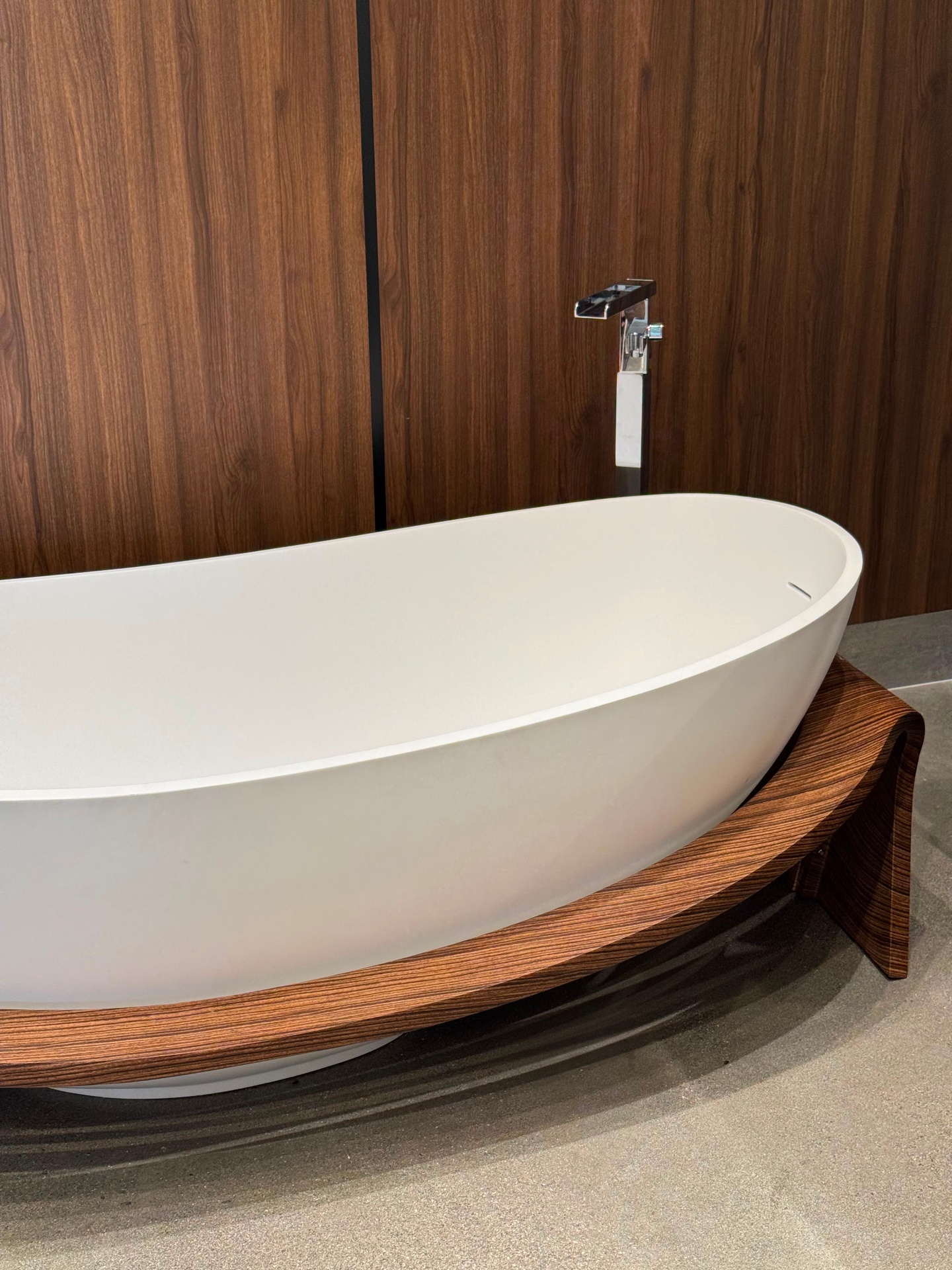 White solid-surface freestanding soaking tub with curved walnut wood base and chrome floor-mount faucet at Ward Design Center bathroom display Kapolei